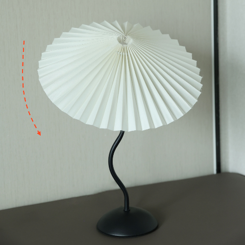 Homira pleated lamp with white pleated lampshade on a black curved stand against a neutral background