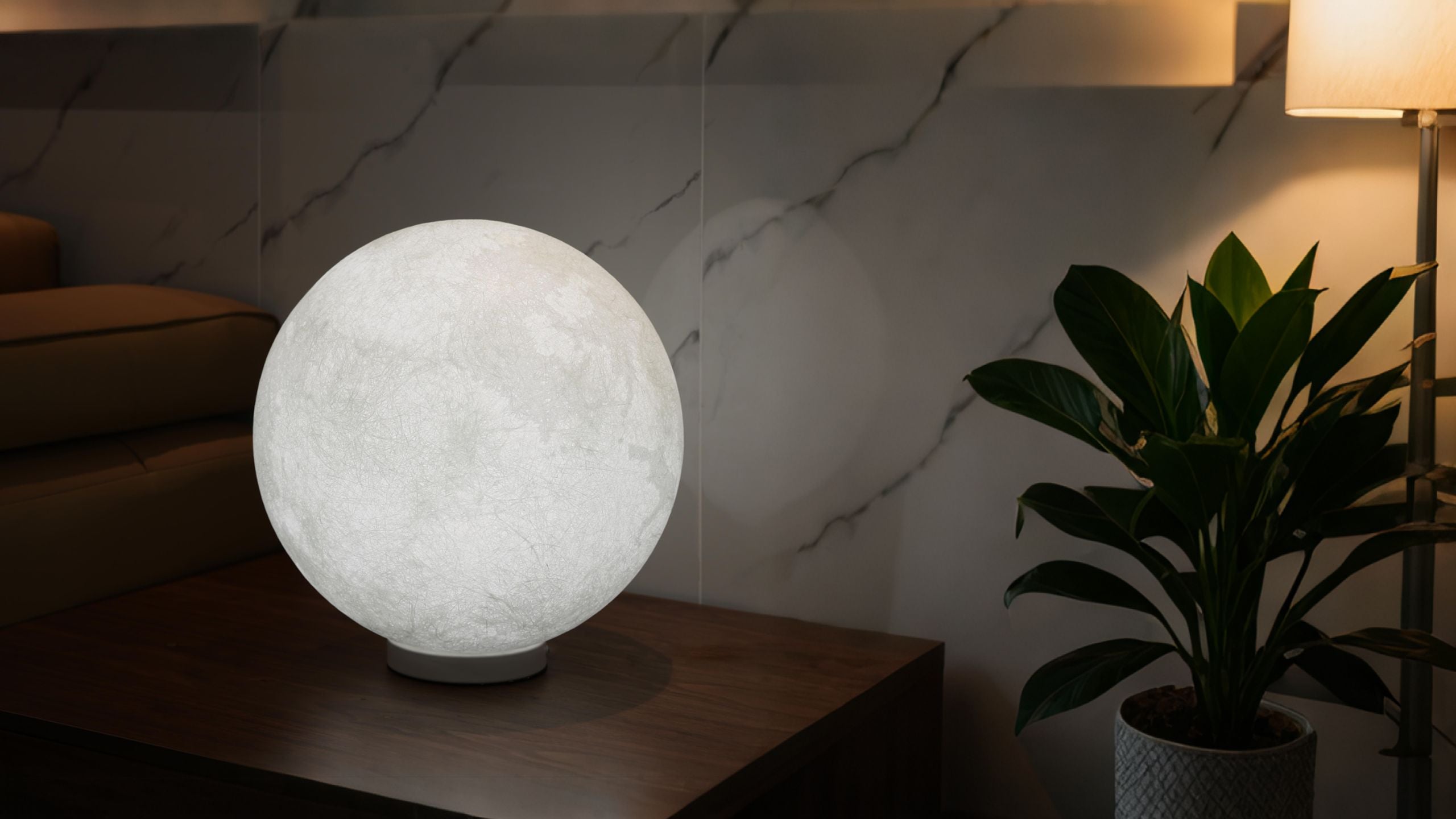 Homira moon-shaped lamp on a table with a plant and marble wall in the background