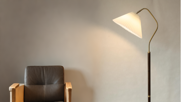 A modern pleated shade floor lamp casts light on a wall next to a leather armchair.